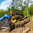 Blue tracked skid steer loader moving large fallen tree logs on rocky terrain with green forest background