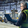 Construction site professional in safety vest using laptop near construction machinery