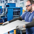 Factory worker wearing safety glasses and protective gloves inspects material at workstation in industrial manufacturing facility