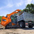 Orange tracked wood chipper loading mulched wood debris into gray dump truck on construction site