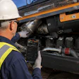 Construction worker in safety gear inspecting engine components of orange industrial truck