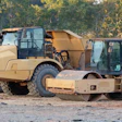 Heavy construction equipment including tan asphalt rollers and compactors lined up at a work site with autumn foliage in background