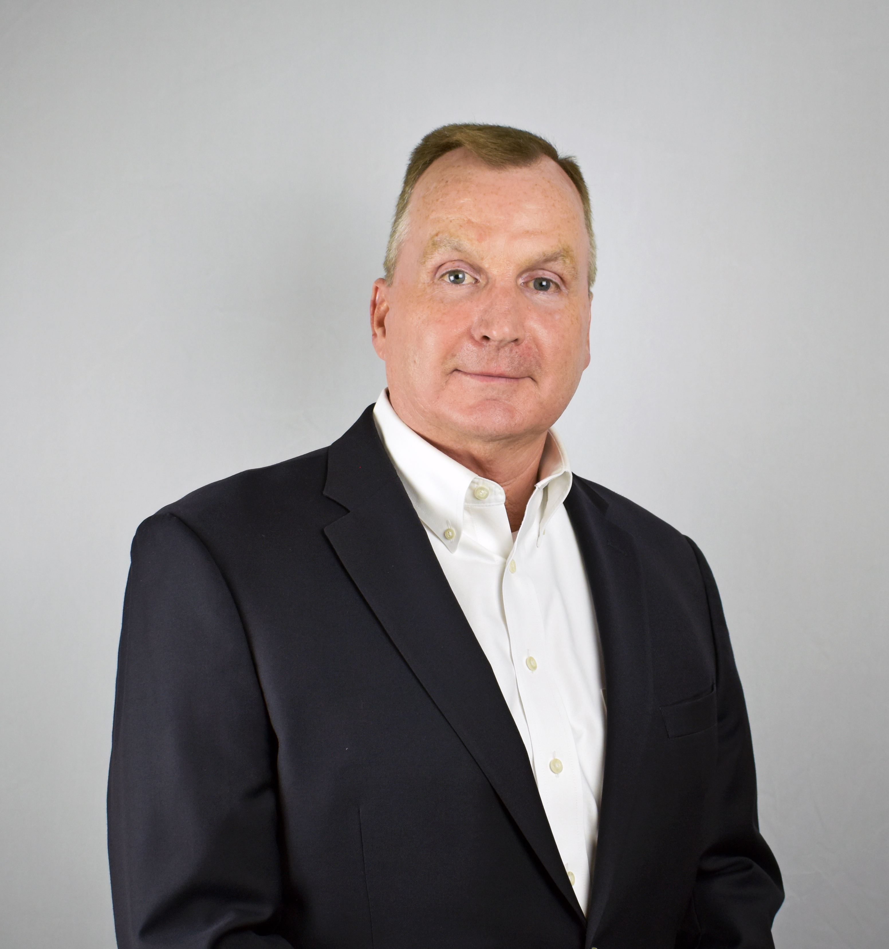 Professional headshot of a middle-aged man in dark blazer and white shirt against neutral background Headshot