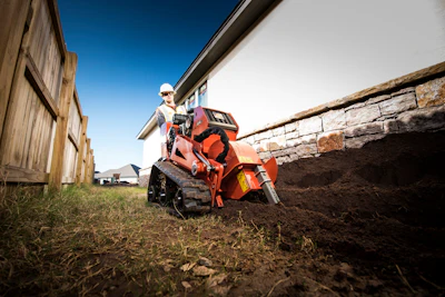 Person operating an orange ATV near farm buildings and stone wall on dirt ground