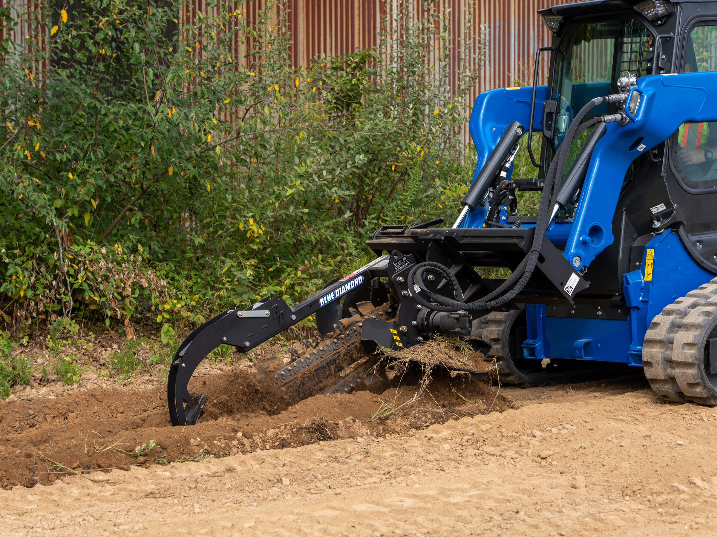Blue compact track loader with grapple bucket clearing brush and debris from dirt ground near corrugated metal fence