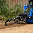 Blue compact track loader with grapple bucket clearing brush and debris from dirt ground near corrugated metal fence