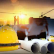 Yellow safety helmet with construction blueprints, ear protection, and workers on building site at sunset