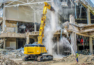 Yellow Kobelco excavator demolishing concrete building with dust spray