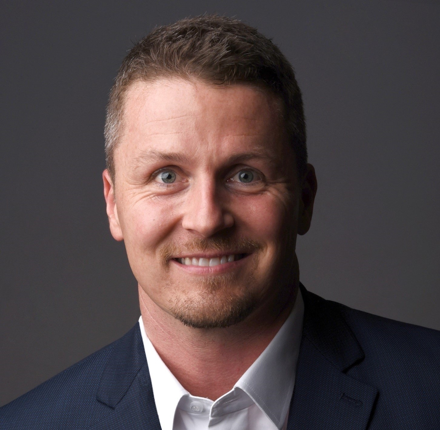 Professional headshot of a smiling man in dark blazer and light shirt against gray background