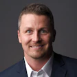 Professional headshot of a smiling man in dark blazer and light shirt against gray background