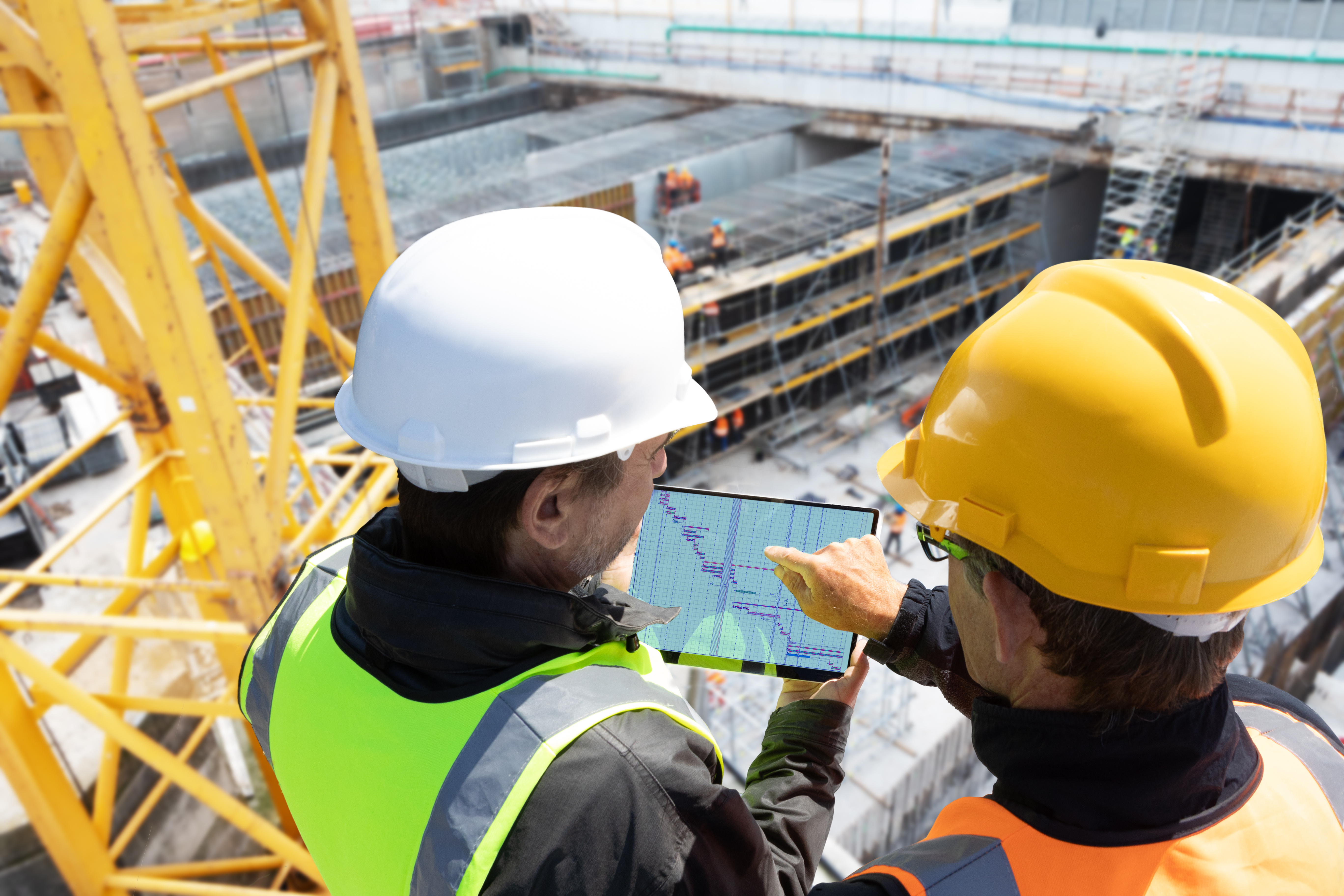 Two construction workers in safety gear reviewing blueprints on a tablet at an active building site.