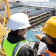 Two construction workers in safety gear reviewing blueprints on a tablet at an active building site.