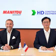 Two business executives at a formal meeting with Manitou Group and HD Construction Equipment logos, French and South Korean flags on table