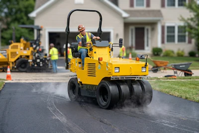 Yellow SAKAI road roller compactor operating on asphalt driveway during residential construction with crew and equipment in background