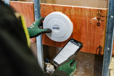 Person in green gloves installing a white WEDGE smart lock on wooden door with smartphone nearby