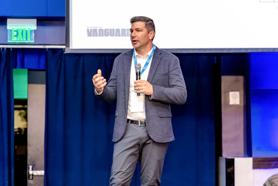 Professional speaker presenting at corporate conference with blue stage backdrop and Vanguard branding