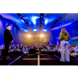 Two business speakers present on stage to an audience in a modern conference hall with blue ambient lighting and warm spotlights.