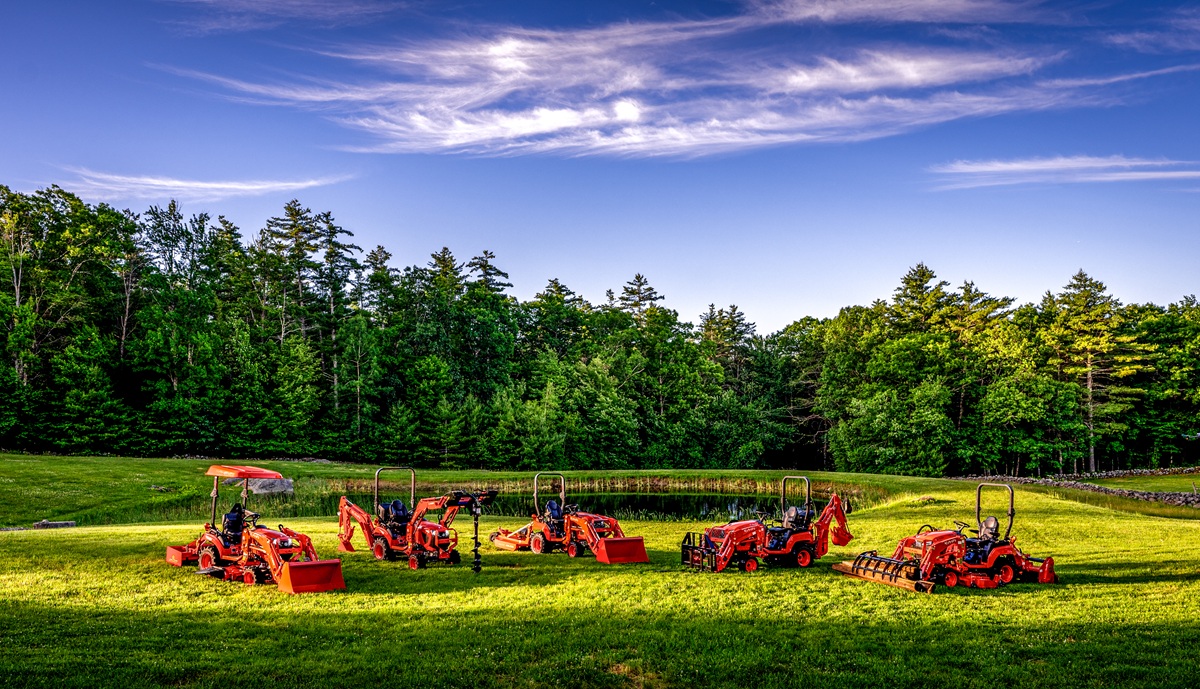 Red farm tractors and equipment lined up in a green field with forest trees under blue sky